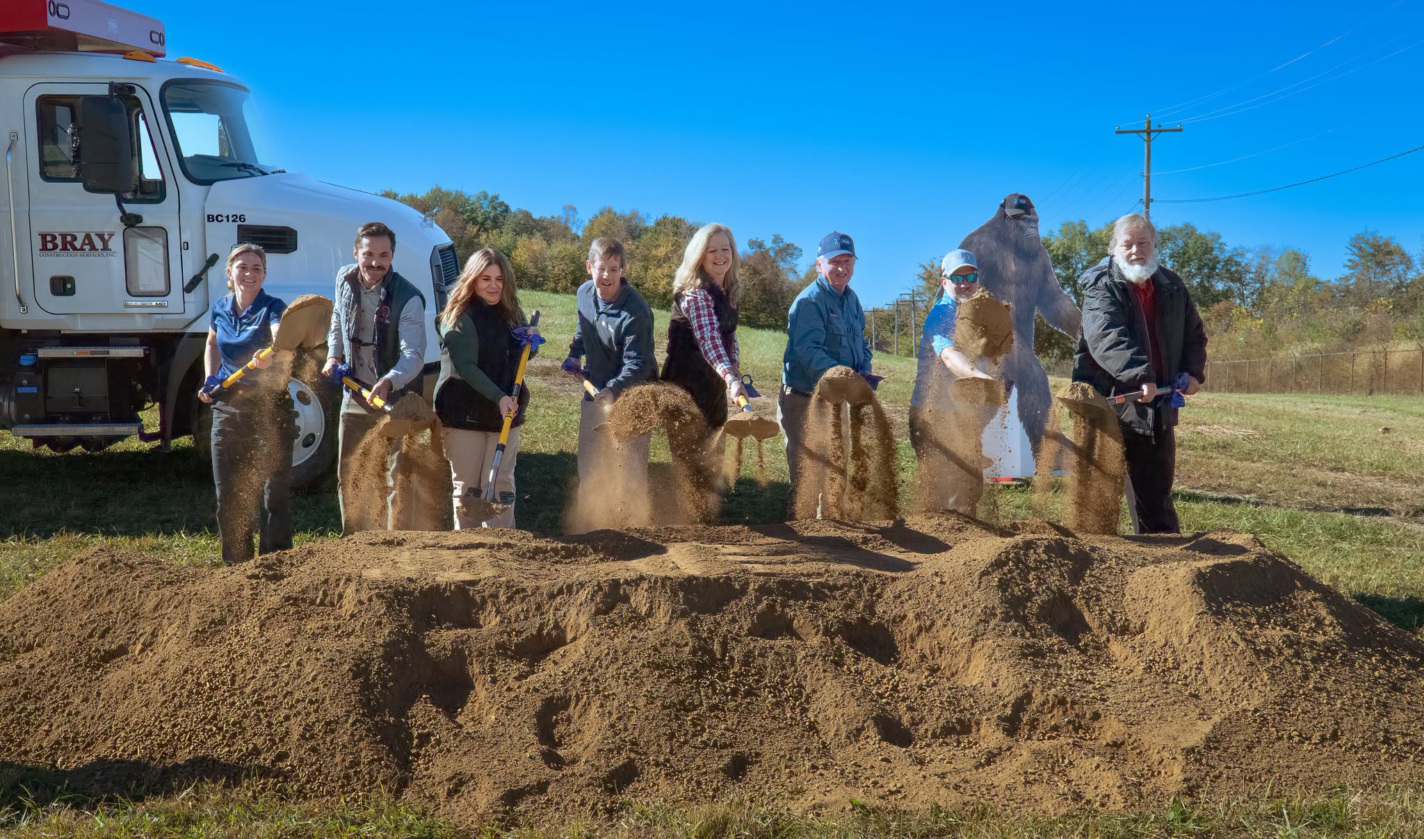 Eight people in a line scooping dirt with shovels. A large truck and a bigfoot cutout are in the background
