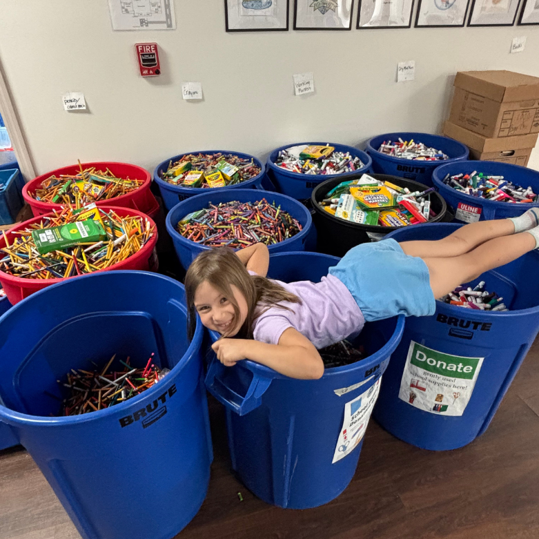 11 large cans full of school supplies such as pencils, markers, and crayons. A young girl lays across the top of them.
